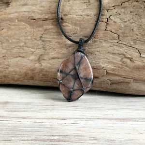 May include: A natural stone pendant necklace. The pendant is a light brown color with dark brown veining, encased in a black woven net. The necklace cord is black and knotted at the top. The background is a piece of weathered wood.