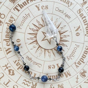 May include: A crystal pendulum with a clear, pointed crystal and a beaded chain featuring blue and black beads. The pendulum rests on a wooden divination board with text including "Yes", "No", and "Check Again".