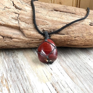 Puede incluir: Un collar con colgante de piedra preciosa roja y cordón negro. La piedra preciosa está encerrada en una red tejida. El collar se exhibe sobre un trozo de madera flotante y una superficie de madera.