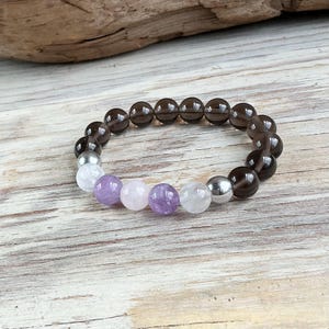 May include: A beaded bracelet featuring a sequence of round, translucent dark brown smoky quartz beads, interspersed with two polished silver-toned metallic beads, two clear quartz beads, and two smooth, round light purple amethyst beads.