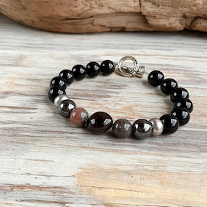 May include: A beaded bracelet featuring a series of smooth, round black beads interspersed with metallic silver-toned beads and two larger, irregular, dark reddish-brown stone beads. The bracelet is secured with a silver-toned toggle clasp.