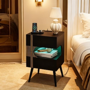 May include: Black nightstand with two drawers and a unique design. The top drawer holds a lamp and a phone charging station. The bottom drawer has a curved opening with a teal interior, displaying books and headphones. The nightstand has gold accents and black legs.