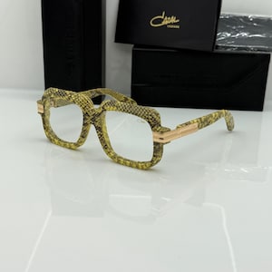 May include: Cazal Legends spectacles featuring a square frame with a yellow and black snakeskin pattern. The temples and bridge are accented with polished gold-tone metal hardware. The brand name Cazal is printed on the lens and the black case.