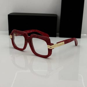May include: Red spectacles with a textured snakeskin pattern on the thick frames. The glasses feature gold-tone metal accents at the hinges and temples, connecting the front rims to the arms. The lenses are clear and the overall design is bold.