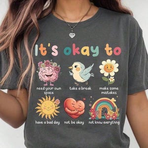 Puede incluir: Camiseta gris oscuro con el texto "It's okay to" sobre ilustraciones coloridas de un cerebro, pájaro, flor, sol, corazón y arcoíris. El diseño incluye las frases "need your own space", "take a break", "make some mistakes", "have a bad day", "not be okay" y "not know everything".