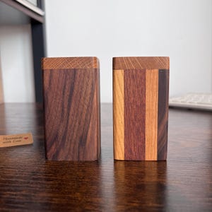 May include: Two wooden rectangular boxes with a dark brown finish. One box is a solid colour, while the other has vertical stripes of varying wood tones. The boxes have a square top and are displayed on a dark wooden surface.
