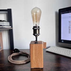 May include: A wooden desk lamp featuring a vintage Edison bulb. The lamp has a rectangular oak base, a black metal stem, and a brass bulb socket. A brown cord and plug are visible. A small tag reads "Handmade with Love".