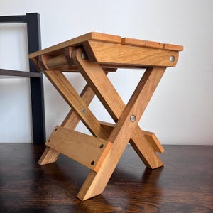 May include: A folding wooden stool with a natural finish. The stool has an X-frame design and a rectangular seat with three slats. The legs are angled for stability and are connected with metal hardware.