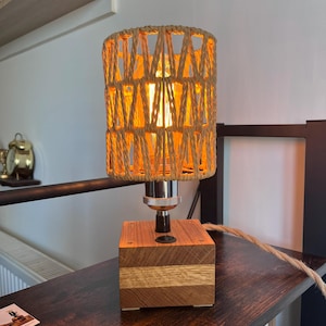 Peut inclure: Lampe de table artisanale avec abat-jour en corde tressée. La lampe présente un socle en bois avec une finition naturelle et un support de lampe chromé. La lumière chaude de l'ampoule traverse l'abat-jour en corde.