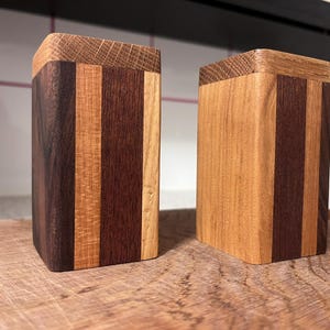 Könnte beinhalten: Zwei Holzkisten mit Deckeln. Die Kisten sind rechteckig und bestehen aus vertikalen Streifen verschiedenfarbigen Holzes, darunter Dunkelbraun, Mittelbraun und Hellbraun. Die Deckel bestehen aus einem helleren Holz mit strukturierter Maserung.