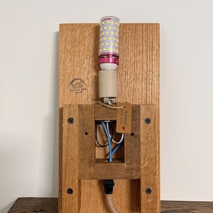 May include: A handmade oak wall sconce with an LED light bulb. The sconce has a rectangular wooden back and a square frame. The light bulb has a pink base. A small tag reads "Handmade with Love".
