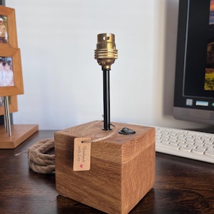 Peut inclure: Une base de lampe en bois faite à la main avec une douille en laiton et une tige noire. La base carrée est en bois brun clair et comporte un petit interrupteur marche/arrêt. Une étiquette "Handmade with Love" est attachée.