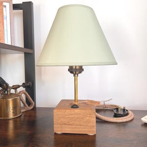 May include: A wooden table lamp with a pale green lampshade. The lamp features a brass stem and a square wooden base. The cord is light brown. The lamp sits on a dark brown wooden surface.
