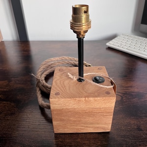 May include: A small wooden lamp base with a black metal stem and a brass bulb socket. The lamp has a black on/off switch and a thick, woven rope cord. The wooden base is square and has a natural wood grain.