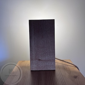 May include: A rectangular wooden lamp with a dark brown, textured grain finish. The lamp emits a soft light from behind its vertical structure and includes a braided fabric power cord extending from the side. A circular logo reads UniqueOakArt.