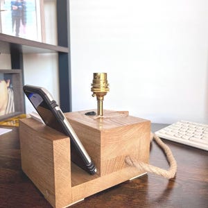 Può includere: Un organizer da scrivania in legno con un supporto per telefono integrato e un portalampada in ottone. L'organizer ha una finitura in legno naturale e un dettaglio in corda. Uno smartphone è posizionato nel supporto per telefono.