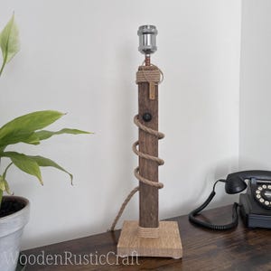 May include: A table lamp featuring a dark brown vertical wooden post mounted on a light-coloured square wooden base. A thick tan rope is wrapped spirally around the post. The lamp includes a silver metal socket, a black power switch, and a small tag.