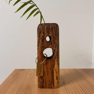 Könnte beinhalten: Eine handgefertigte Holzvase mit rechteckiger Form und zwei kreisförmigen Öffnungen. Ein grüner Pflanzenstiel ist in die Oberseite eingesetzt. Ein kleines Etikett mit der Aufschrift "Handmade with love" ist mit Bindfaden befestigt. Die Vase steht auf einer Holzoberfläche.
