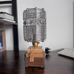 May include: A table lamp with a metal wire lampshade with floral detailing. The lamp sits on a wooden base with a brass fitting and a plug. A small tag reads "Handmade with Love."