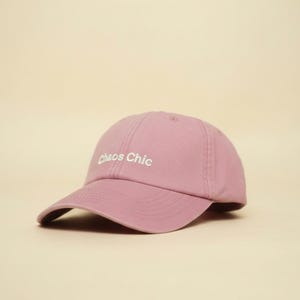 Könnte beinhalten: Staubrosa Baseballkappe mit dem Text "Chaos Chic" in Weiß gestickt. Die Kappe hat einen gebogenen Schirm und ein klassisches Sechs-Panel-Design. Der Hintergrund ist einfarbig hellbeige.