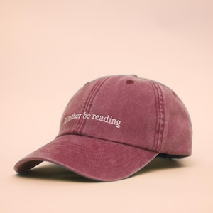 Puede incluir: Gorra de béisbol granate con visera curva. La gorra presenta el texto bordado "I'd rather be reading" en blanco. Tiene un diseño clásico de seis paneles y un cierre de correa de tela.