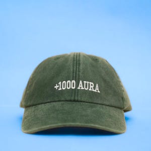 Peut inclure: Casquette de baseball vert fonc&eacute; avec le texte "+1000 AURA" brod&eacute; en blanc. La casquette est faite d'une mati&egrave;re douce et poss&egrave;de une visi&egrave;re incurv&eacute;e. Le fond est bleu clair uni.
