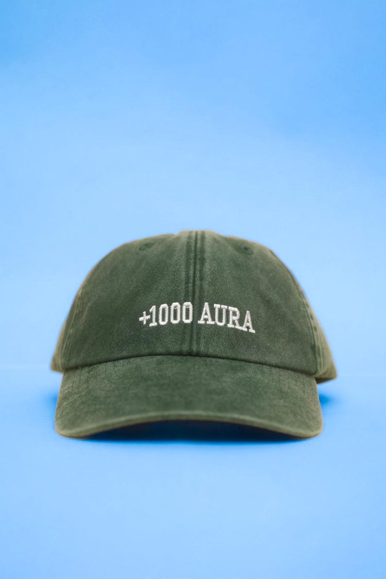 Peut inclure: Casquette de baseball vert fonc&eacute; avec le texte "+1000 AURA" brod&eacute; en blanc. La casquette est faite d'une mati&egrave;re douce et poss&egrave;de une visi&egrave;re incurv&eacute;e. Le fond est bleu clair uni.