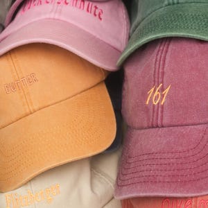 Könnte beinhalten: Eine Sammlung von Baseballkappen in verschiedenen Farben, darunter Rosa, Orange, Grün und Burgunder. Jede Kappe hat gestickte Texte oder Zahlen, wie "Butter", "161" und "rave to the grave". Die Kappen sind gestapelt und zeigen ihre unterschiedlichen Designs.