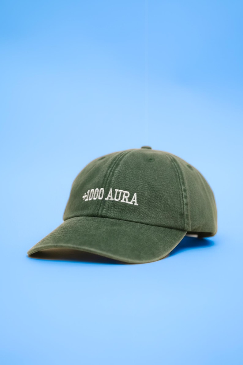 Peut inclure: Casquette de baseball vert olive avec le texte "+1000 AURA" brod&eacute; en blanc. La casquette a une visi&egrave;re incurv&eacute;e et une conception classique &agrave; six panneaux. Le fond est bleu clair uni.