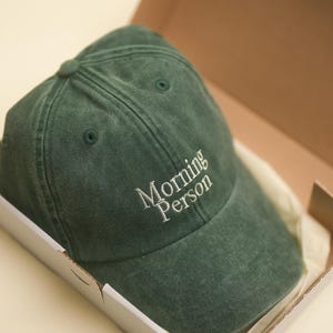 Peut inclure: Casquette de baseball vert fonc&eacute; avec le texte brod&eacute; "Morning Person" en blanc. La casquette est &agrave; l'int&eacute;rieur d'une bo&icirc;te en carton blanche avec un int&eacute;rieur beige.