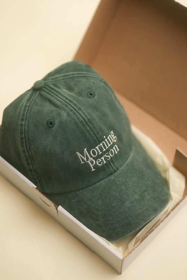 Peut inclure: Casquette de baseball vert fonc&eacute; avec le texte brod&eacute; "Morning Person" en blanc. La casquette est &agrave; l'int&eacute;rieur d'une bo&icirc;te en carton blanche avec un int&eacute;rieur beige.