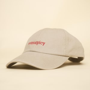 Könnte beinhalten: Eine beige Baseballkappe mit dem Wort neurospicy, das in rotem Garn auf das Vorderteil gestickt ist. Die Kappe hat einen gebogenen Schirm und eine strukturierte Krone mit sichtbaren Nähten entlang der Paneele, was ein schlichtes Design ergibt.