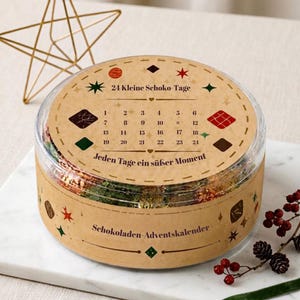 May include: A round, clear plastic container holding a chocolate advent calendar with a tan paper label. The label features the text 24 Kleine Schoko-Tage, Jeden Tage ein süßer Moment, and Schokoladen-Adventskalender, along with numbers 1 through 24 and colorful star and diamond icons.