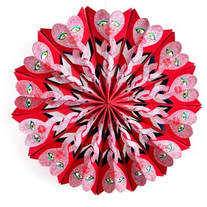 Könnte beinhalten: Ein roter Papierfächer mit einem herzförmigen Design mit glitzernden rosa Gesichtern mit grünen Augen und roten Lippen. Der Fächer ist an einer weißen Schnur aufgehängt und bildet ein dekoratives, auffälliges Stück.