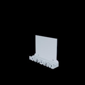 Op de afbeelding: Een witte wandplank met de tekst "NOW PLAYING" in 3D-lettertype. De plank heeft een rechthoekig achterpaneel en een kleine richel. Twee kleine gaten zijn zichtbaar op het achterpaneel.