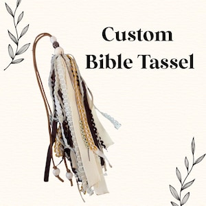 May include: A custom Bible tassel with a mix of ribbons in cream, brown, and gold. The tassel includes lace and beaded accents, and the text "REDEEMED" is spelled out on a strand. The tassel is attached to a leather cord.