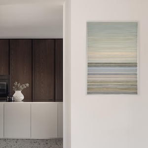 Op de afbeelding: Abstract kunstwerk met horizontale lijnen in blauw, grijs en geel. Het rechthoekige stuk meet 56 x 80 cm. De afbeelding toont ook een moderne keuken met donkere houten kasten en een witte vaas met bloemen, gezien door een deuropening.