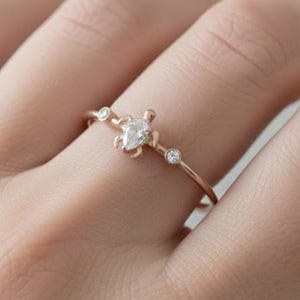 May include: A delicate rose gold ring featuring a pear-shaped diamond center stone and two smaller round diamonds. The ring is set on a thin band, creating a minimalist and elegant design. The ring is shown on a hand.