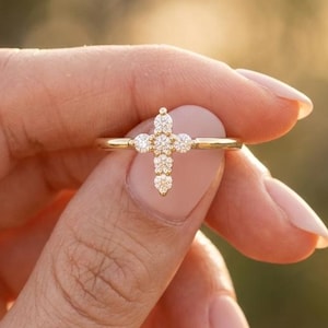 May include: A gold ring featuring a cross-shaped centrepiece composed of small, round, clear stones. The band is thin and polished, with the cross design set horizontally across the finger, showcasing a delicate and symmetrical arrangement.