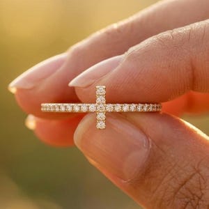 May include: A thin gold ring featuring a horizontal band encrusted with small clear stones and a vertical cross-shaped accent also set with small clear stones. The metal has a warm yellow tone and the stones provide a bright, reflective sparkle.