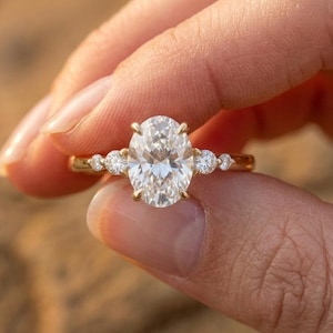 Puede incluir: Un anillo de oro con una gran piedra central de corte ovalado sujeta por cuatro garras, flanqueada por dos piedras más pequeñas de corte redondo a cada lado. El aro es de oro amarillo pulido, con un total de cinco piedras transparentes.