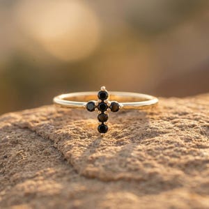 Puede incluir: Un anillo de oro con un adorno en forma de cruz compuesto por cinco pequeñas piedras preciosas negras redondas. La banda es fina y pulida, con el diseño de cruz fijado de forma segura en la parte superior, mostrando el contraste entre el oro y las piedras.