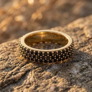 May include: A gold band ring featuring three rows of small, round, black stones set closely together around the entire circumference of the metal band, creating a textured, pavé-style surface with a polished gold finish on the interior.