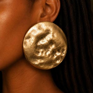 May include: A large, circular earring with a matte gold finish and a textured, hammered surface. The earring features an irregular, organic shape with deep indentations and raised areas across its face, creating a distinct, uneven appearance.