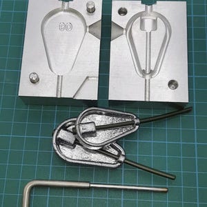 May include: A metal fishing lure mold with two halves, a finished lure, and a tool. The mold is silver and has a teardrop shape with the number "80" engraved. The finished lure is also silver with a wire extending from it.