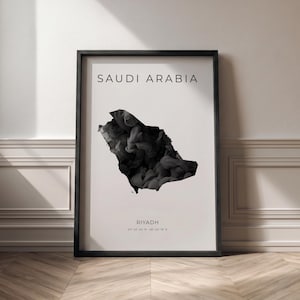 Op de afbeelding: Een ingelijste kunstprint met een zwart-wit kaart van Saoedi-Arabië. De omtrek van het land is gevuld met een rokerig, abstract ontwerp. De tekst "SAUDI ARABIA" staat bovenaan en "RIYADH" met coördinaten onderaan.