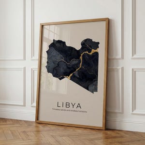 May include: Framed art print featuring a map of Libya in dark blue with gold accents. The print includes the country's name and the phrase "Timeless sands and endless horizons" in a minimalist style. The frame is made of light-colored wood.