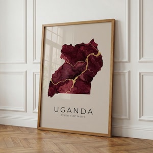 Könnte beinhalten: Gerahmter Kunstdruck mit einer Karte von Uganda in Burgundertönen mit goldenen Akzenten. Das Wort "UGANDA" ist darunter gedruckt, zusammen mit den Koordinaten 0°18' N 32°34' E. Der Druck ist in einem hellen Holzrahmen.