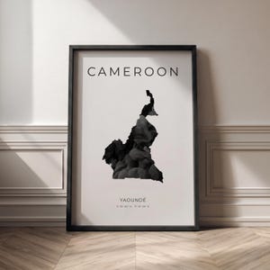 May include: A framed print featuring a black and white map of Cameroon. The word "CAMEROON" is at the top, with "YAOUNDE" and coordinates below the map. The map is filled with a dark, textured design, set against a white background.