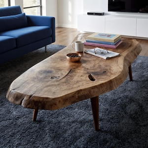 May include: A live edge wooden coffee table with a natural, organic shape. The table has a light brown finish and sits on four tapered legs. A blue sofa and a gray rug are in the background.